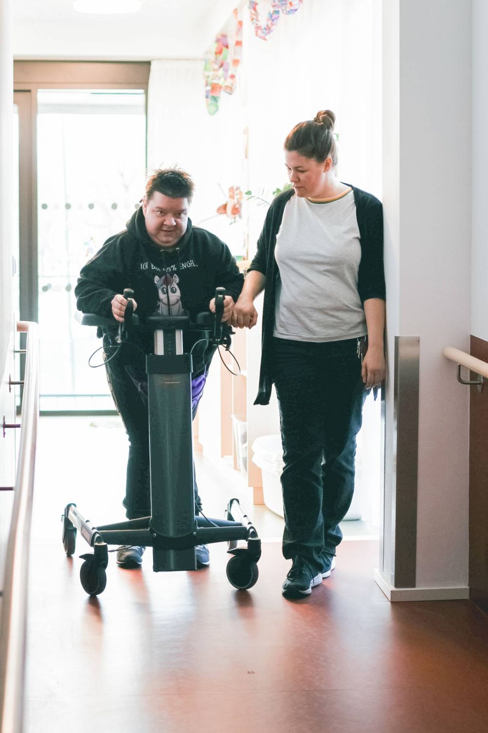 eine Frau mit Gehbehinderung übt mit einem Gehtrainer; die Unterstützerin geht neben ihr