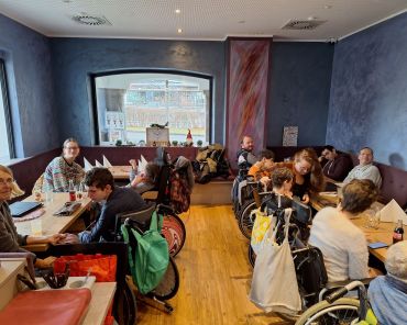 eine Gruppe von Menschen mit Behinderungen in Rollstühlen und Unterstützer_innen in einem Cafè an mehreren Tischen