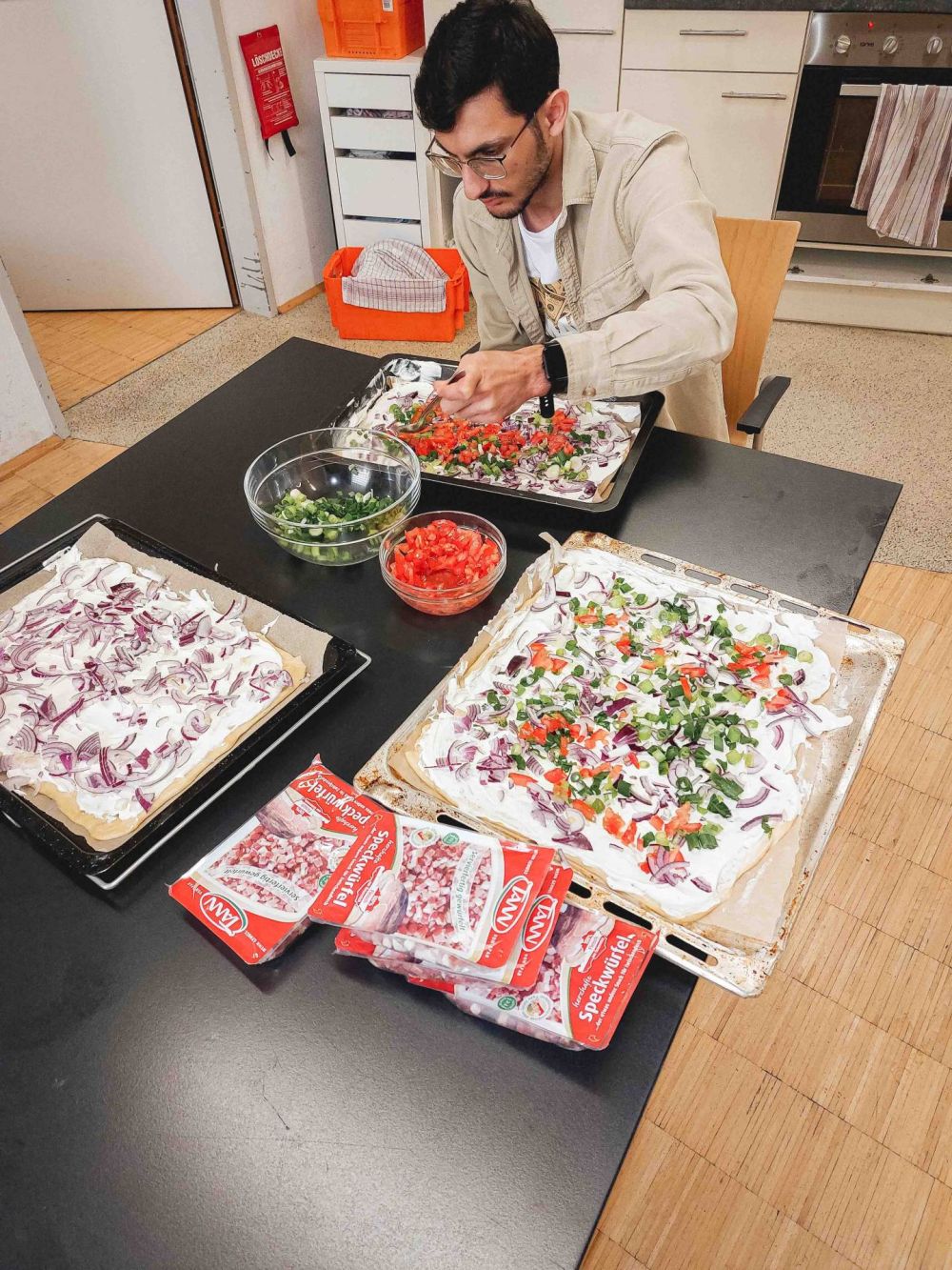 ein junger Mann mit schwarzen Haaren sitzt an einem Tisch, vor ihm liegen drei Blech mit ungebackenem Flammkuchen und zwei Schüsseln mit Zutaten für den Flammkuchen-Belag