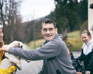 ein Mann streichelt einen kleinen Hund, er lächelt; eine Frau mit gelber Jacke hält den Hund und lächelt ebenfalls