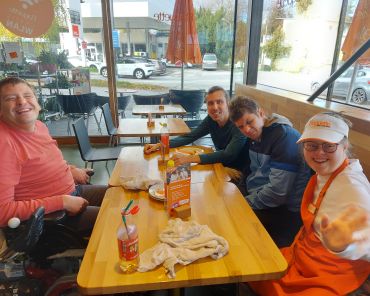 drei Männer und eine Frau in Bäckersschürze sitzen um einen Tisch und grinsen in die Kamera