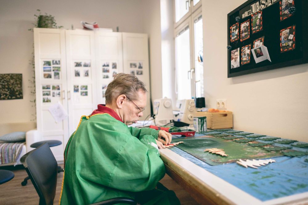ein Mann mit einer grünen Malerschürze bemalt kleine Weihnachtsbäume aus Holz