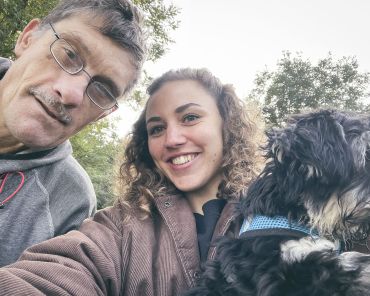 ein Mann mit Brille und eine junge Frau mit Locken schauen in die Kamera und machen ein Selfie; die Frau trägt einen kleinen Hund im Arm