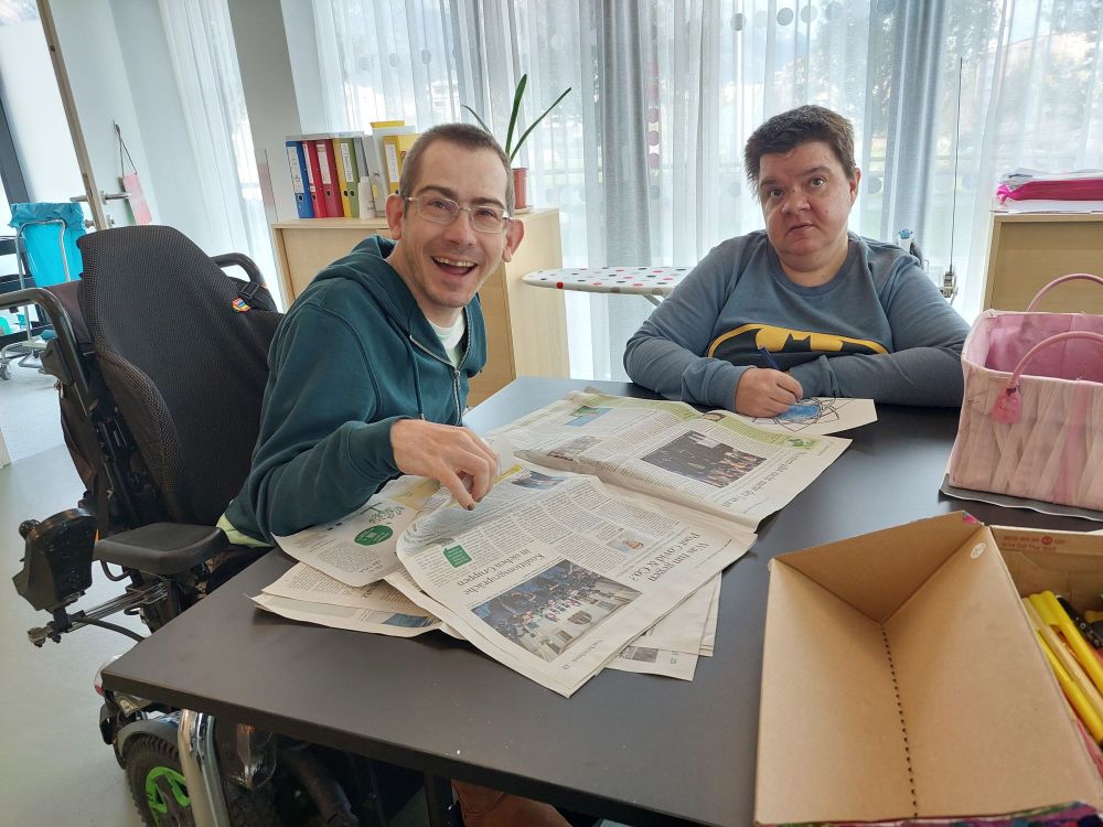 zwei Menschen mit Behinderungen sitzen an einem Tisch und lesen eine Zeitung