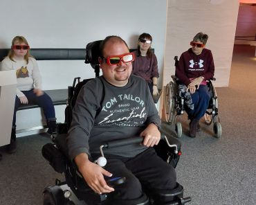 mehrere Personen in einem abgedunkelten Raum mit 3D Brillen auf; zwei Personen im Rollstuhl