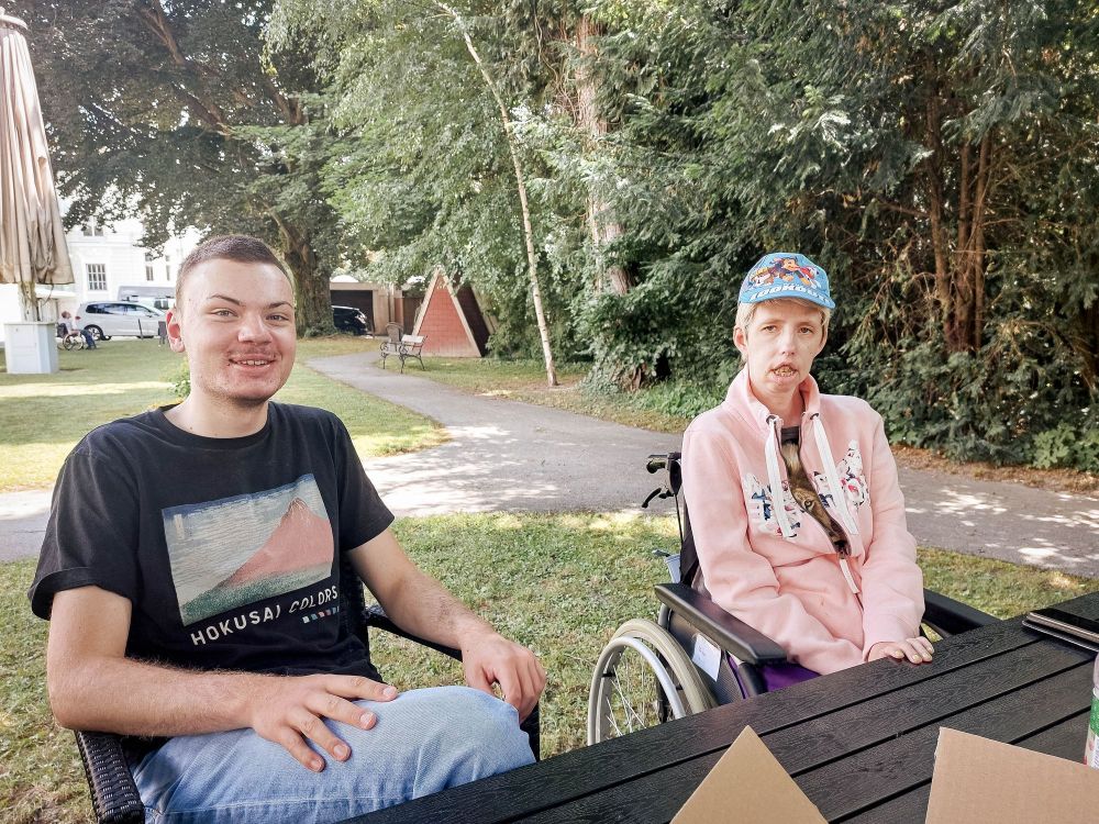 eine Frau im Rollstuhl sitzt an einem Tisch, ein junger Mann sitzt daneben und lächelt; die beiden schauen in die Kamera