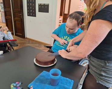 ein Mann mit Behinderung schneidet mit einer Unterstützungsperson einen Schokokuchen an