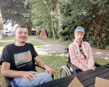 eine Frau im Rollstuhl sitzt an einem Tisch, ein junger Mann sitzt daneben und lächelt; die beiden schauen in die Kamera