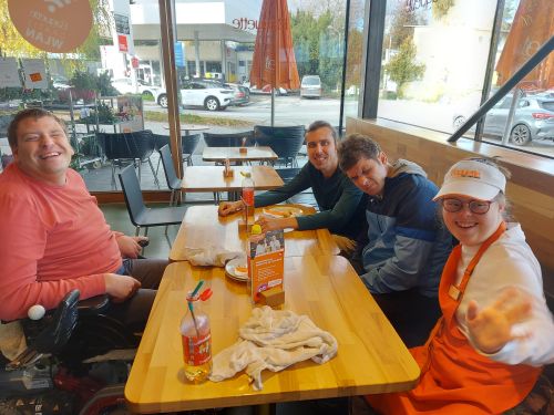 drei Männer und eine Frau in Bäckersschürze sitzen um einen Tisch und grinsen in die Kamera