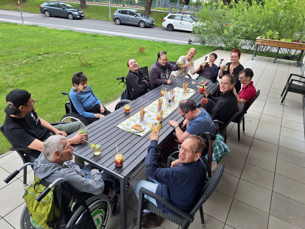 eine Gruppe von Menschen mit Behinderungen sitzen um einen Tisch auf der Terrasse; vor ihnen stehen alkoholfreie Cocktails