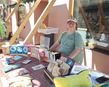 ein Mann mit Downsyndrom steht vor einem Marktstand mit verschiedenen Produkten und lächelt in die Kamera