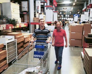 zwei Männer mit Behinderung schieben einen Einkaufswagen durch das Lager eines Möbelhauses