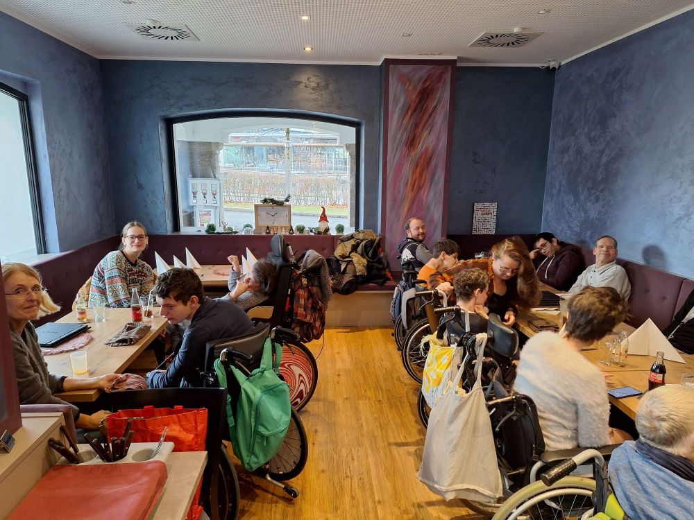 eine Gruppe von Menschen mit Behinderungen in Rollstühlen und Unterstützer_innen in einem Cafè an mehreren Tischen