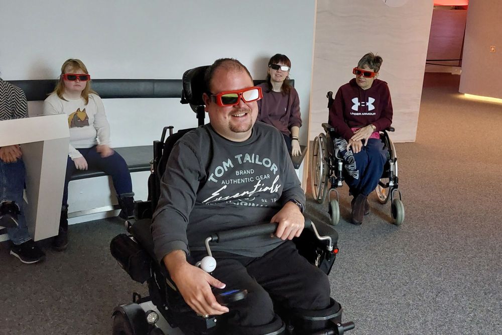 mehrere Personen in einem abgedunkelten Raum mit 3D Brillen auf; zwei Personen im Rollstuhl