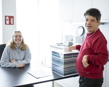 ein Mann mit Behinderung lächelt in die Kamera, er steckt einen Zettel in eine Wahlurne, dahinter sitzt eine junge Frau an einem Tisch