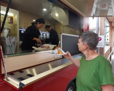 eine Frau erhält einen Teller mit Burger und Pommes aus einem roten Foodtruck; darin arbeiten eine Frau und ein Mann, die das Essen zubereiten