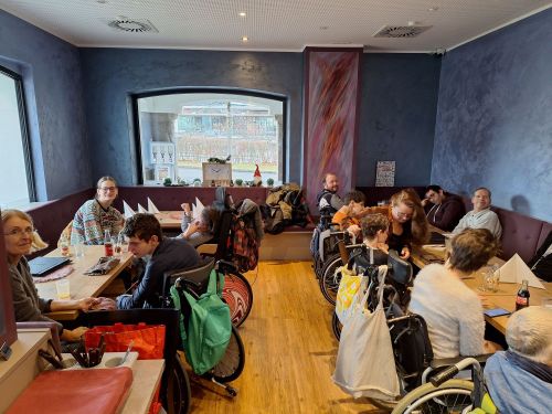 eine Gruppe von Menschen mit Behinderungen in Rollstühlen und Unterstützer_innen in einem Cafè an mehreren Tischen
