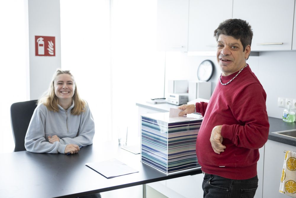 ein Mann mit Behinderung lächelt in die Kamera, er steckt einen Zettel in eine Wahlurne, dahinter sitzt eine junge Frau an einem Tisch