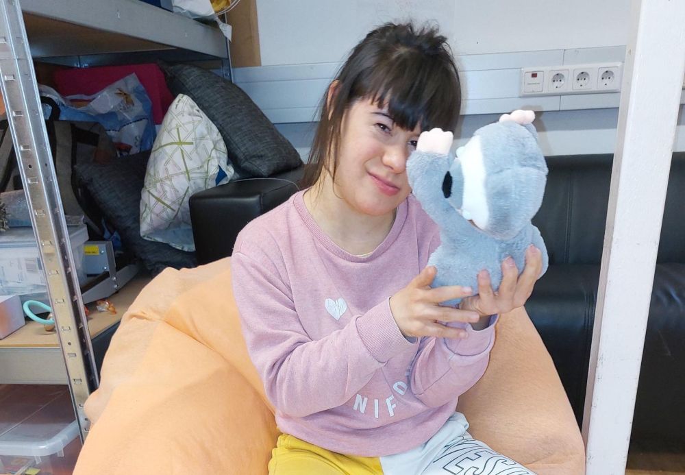 eine junge Frau mit Behinderung sitzt in einem Sitzsack und hält ein graues Stofftier hoch