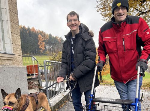 zwei Männer stehen im Freien, einer führt einen Hund mit Maulkorb an der Leine, der andere geht mit einem Rollator; beide lächeln in die Kamera