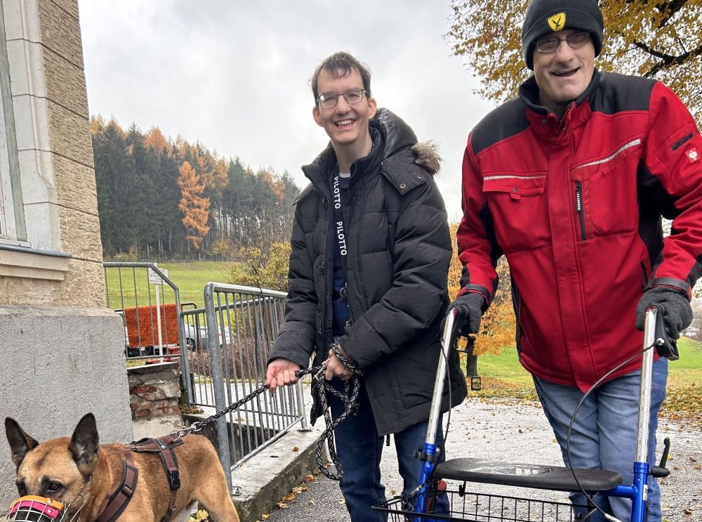 zwei Männer stehen im Freien, einer führt einen Hund mit Maulkorb an der Leine, der andere geht mit einem Rollator; beide lächeln in die Kamera