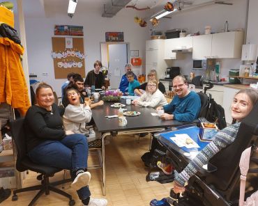 eine Gruppe Menschen mit und ohne Behinderungen sitzen um einen Tisch herum und lächeln in die Kamera