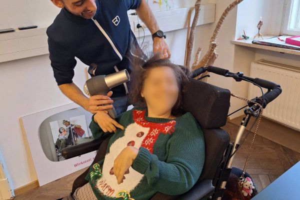 Eine Frau im Rollstuhl hält einen Föhn an ihre Haare, ein Unterstützer steht daneben und hilft ihr