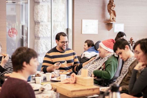 viele Menschen sind im Raum; ein junger Mann gibt einem Mann im Rollstuhl mit Weihnachtsmütze Essen ein