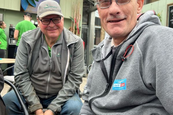 zwei Männer sitzen nebeneinander; im Hintergrund ein Glasdach und Gärtnerei-Mitarbeiter mit grünen Shirts