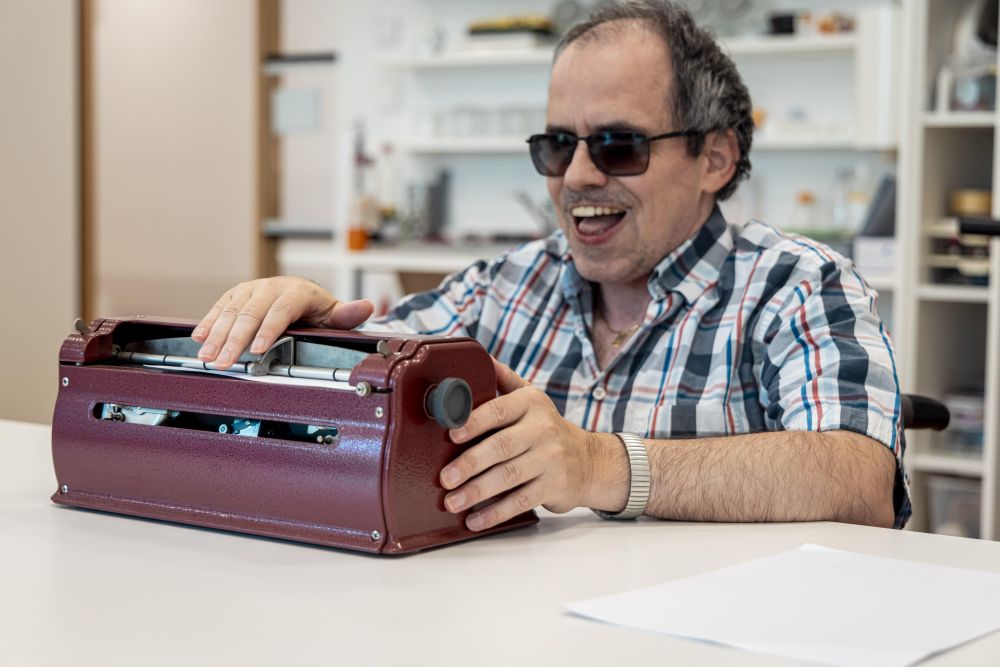 Ein Mann mit Sehbehinderung sitzt an einem Tisch und bedient eine Braille-Schreibmaschine. Er trägt eine Sonnenbrille und lächelt.