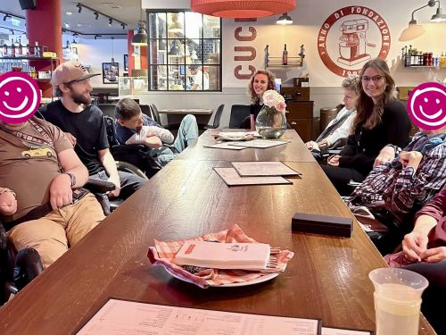 eine Gruppe Erwachsener sitzt um einen Tisch in einer Pizzeria; manche schauen in die Kamera