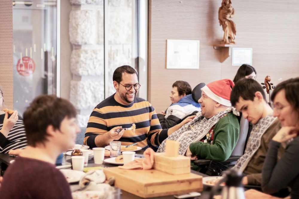 viele Menschen sind im Raum; ein junger Mann gibt einem Mann im Rollstuhl mit Weihnachtsmütze Essen ein
