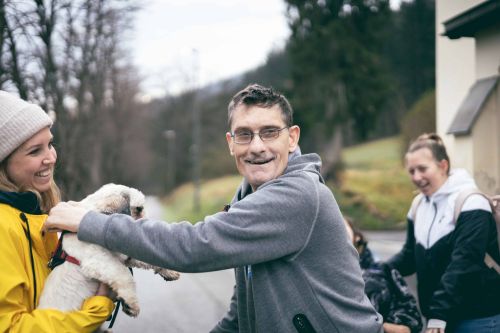 ein Mann streichelt einen kleinen Hund, er lächelt; eine Frau mit gelber Jacke hält den Hund und lächelt ebenfalls