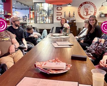 eine Gruppe Erwachsener sitzt um einen Tisch in einer Pizzeria; manche schauen in die Kamera