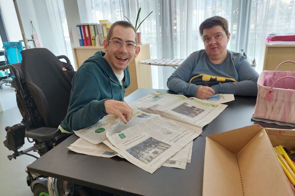 zwei Menschen mit Behinderungen sitzen an einem Tisch und lesen eine Zeitung