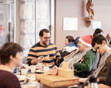 viele Menschen sind im Raum; ein junger Mann gibt einem Mann im Rollstuhl mit Weihnachtsmütze Essen ein