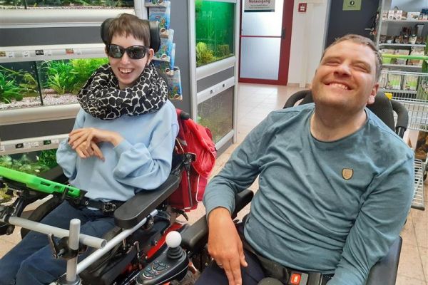eine Frau und ein Mann mit Behinderung im Rollstuhl in einem Geschäft; beide lächeln in die Kamera