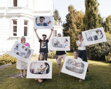 sechs Erwachsene stehen auf einer Wiese und halten Tafeln mit Bildern in die Luft
