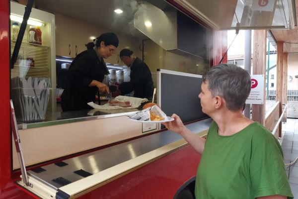 eine Frau erhält einen Teller mit Burger und Pommes aus einem roten Foodtruck; darin arbeiten eine Frau und ein Mann, die das Essen zubereiten