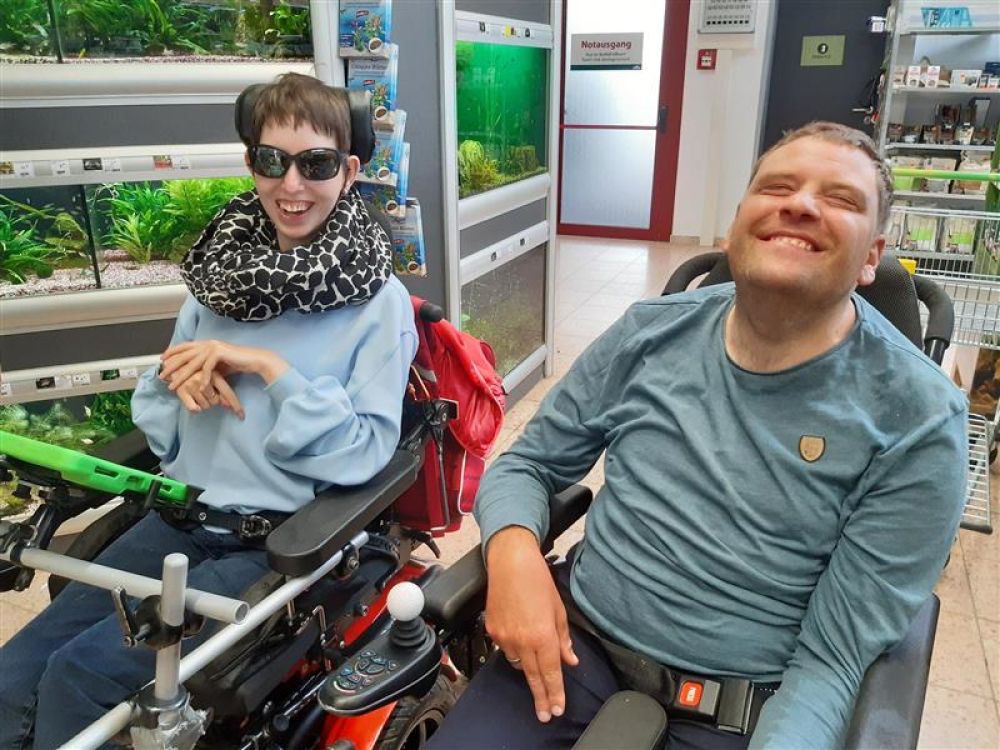 eine Frau und ein Mann mit Behinderung im Rollstuhl in einem Geschäft; beide lächeln in die Kamera