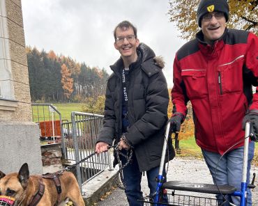 zwei Männer stehen im Freien, einer führt einen Hund mit Maulkorb an der Leine, der andere geht mit einem Rollator; beide lächeln in die Kamera