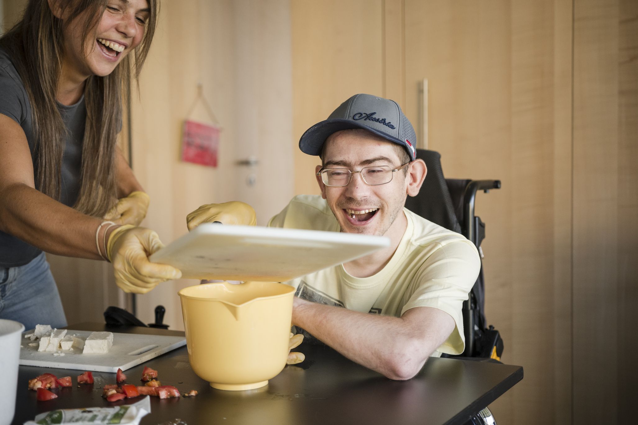 ein Mann mit Behinderung im Rollstuhl schiebt geschnittenes Gemüse von einem Schendiebrett in eine Schüssel; eine lächelnde Betreuerin hält das Brett