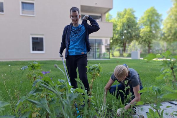 Ein Mann mit Behinderung und eine Unterstützerin arbeiten im Garten einer Wohnanlage an einem grünen Beet